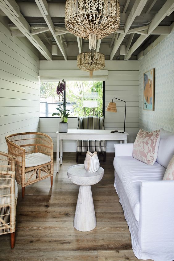 a white shed home office with planked walls, a white sofa, rattan chairs, a white desk and a striped chair plus bead chandeliers