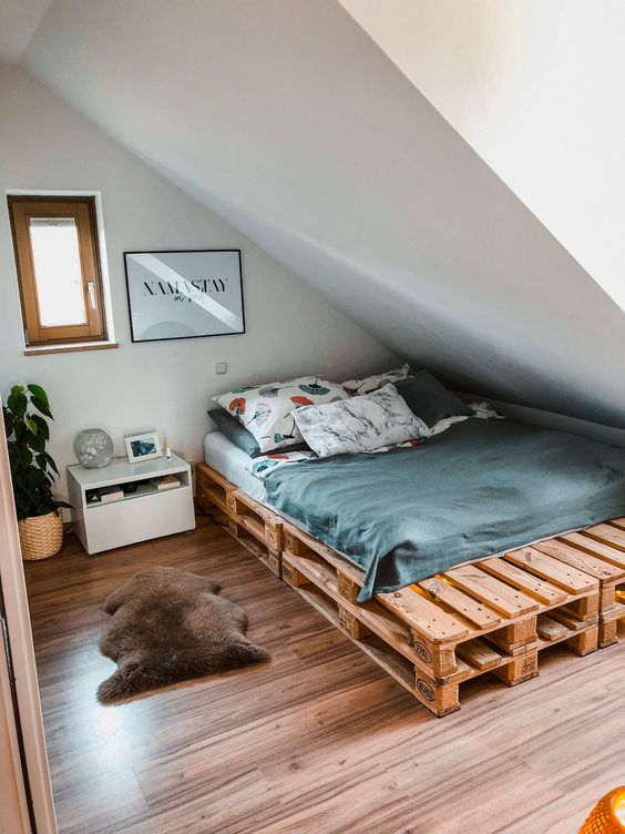 an attic bedroom with a pallet bed and grey bedding, a nightstand, artwork and a potted plant