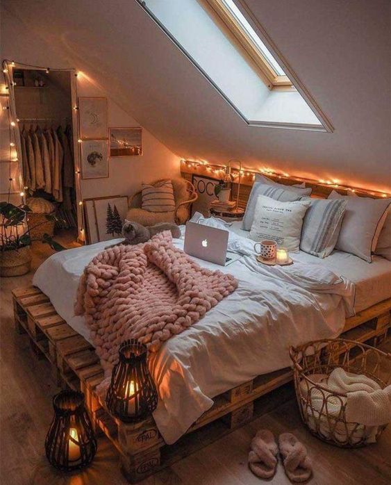 an attic bedroom with a pallet bed with neutral bedding, a rattan chair with pillows, lights and potted plants