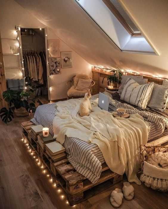 an attic bedroom with a pallet bed with printed bedding and lights, potted greenery and a basket with pillows