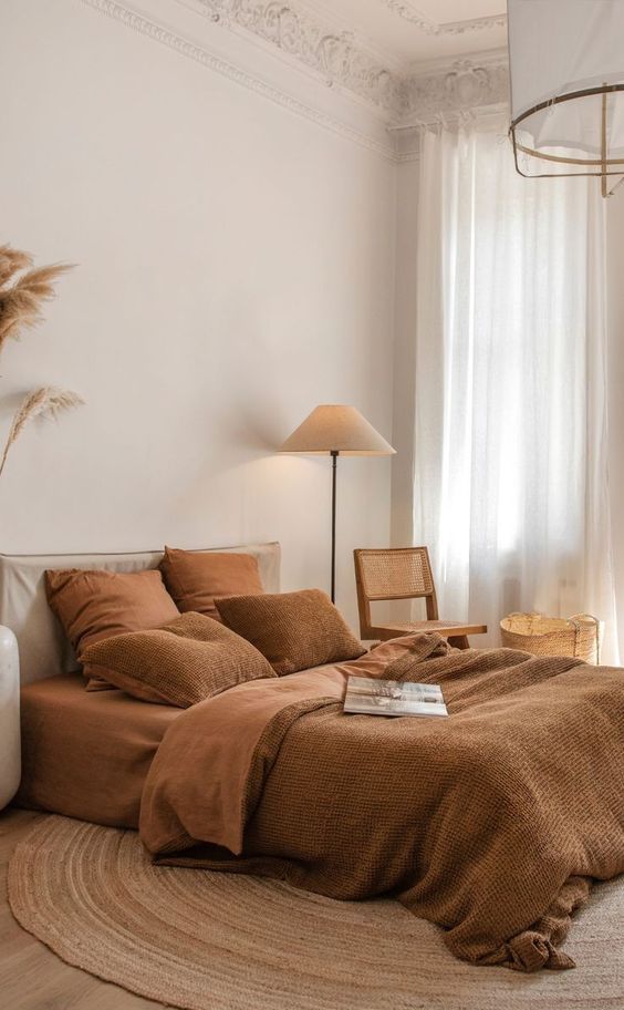 an earthy bedroom with rust bedding, a jute rug, a cane chair, a floor lamp and some pampas grass for a boho feel