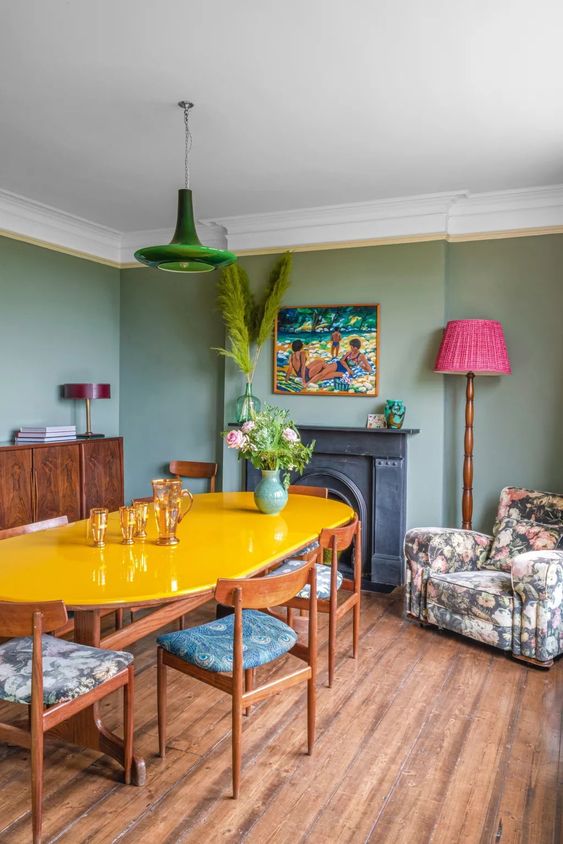 an eclectic dining room with green walls, a fireplace, a yellow table, a pink floor lamp, a green pendant lamp and a floral chair