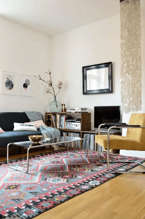 an eclectic living room with a bold boho rug, a black sofa with pastel pillows, a yellow chair, a coffee table and a bookshelf with vinyl