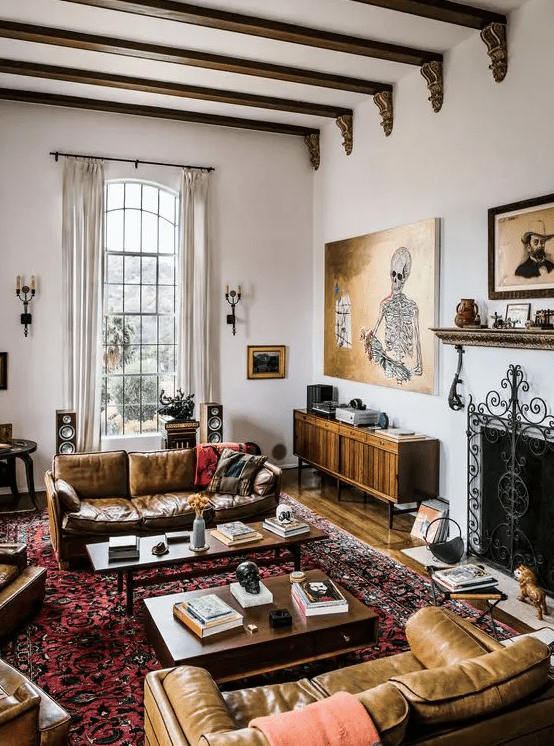 an eclectic living room with a fireplace, beige leather sofas, coffee tables, a printed rug, colorful pillows, artwork and wooden beams