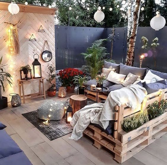 an eye-catching terrace with a pallet corner sofa and pillows, some tables and rugs, candle lanterns, lights and greenery