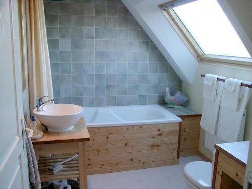 a small attic bathroom clad with grey tiles, with a skylight, a bathtub clad with wood and wooden furniture for a natural touch