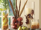 a cool fall centerpiece of glasses with faux fruit, feathers, dried leaves, nuts and acorns is very natural