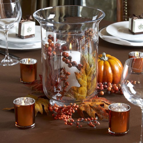 a fall wedding centerpiece of a tall glass vase with a candle, berries and a leaf and candles around it