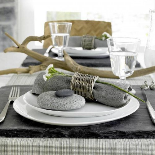 a natural coastal setting with a grey napkin with a wicker napkin ring, some pebbles and driftwood as a centerpiece