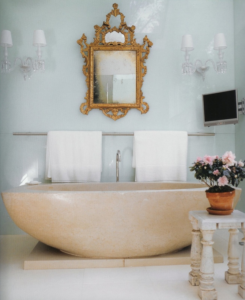 a refined bathroom with an oval stone tub, chic wall sconces, an ornate mirror and potted blooms looks glam and bright