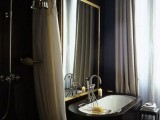 a contemporary dark bathroom with black walls, a black stone clad tub, a gold frame mirror and curtains for the shower space