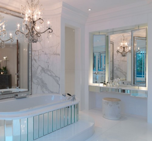 a luxurious bathroom in white marble, with a crystal chandelier, a mirror clad bathtub and a mirror over it for a statement