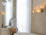 a contemporary bathroom in neutrals, a crystal chandelier, a mirror wall, an oval tub, pillar candles on the shelf over it