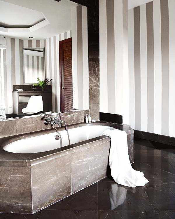 a modern luxurious bathroom with striped walls, a dark floor, a stone bathtub and a whole mirror wall over it to enlarge the space